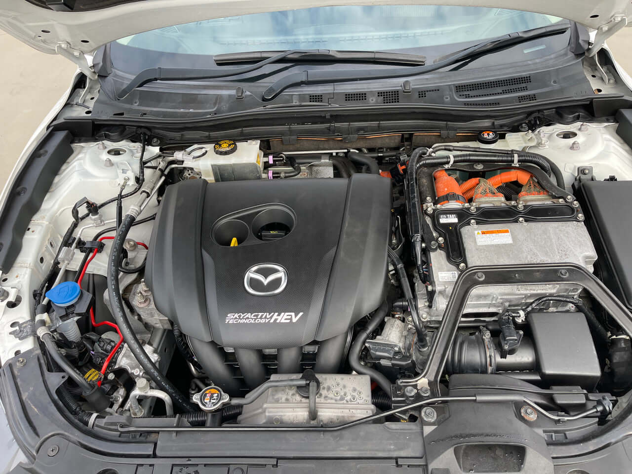 Engine compartment of the 2014 Mazda Axela 2.0L Hybrid showing hybrid components and Mazda branding.