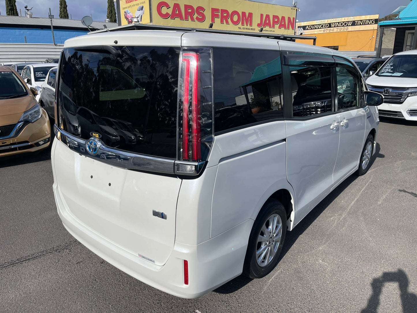 2018 Toyota Noah 1.8L Hybrid G Edition 7 seater rear view with clear sky and dealership in the background.