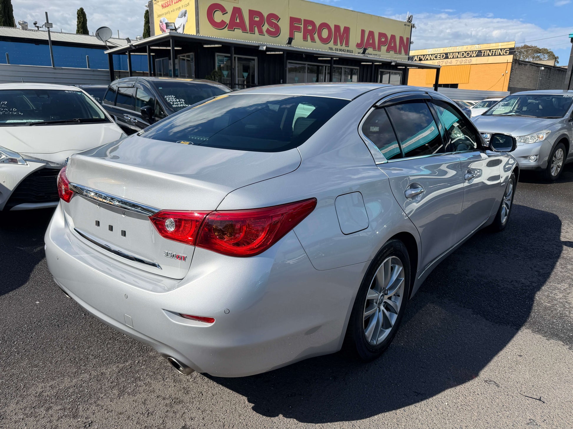 2014 Nissan Skyline V6 3.5L Hybrid parked at a car dealership showing its sleek silver design and rear profile.